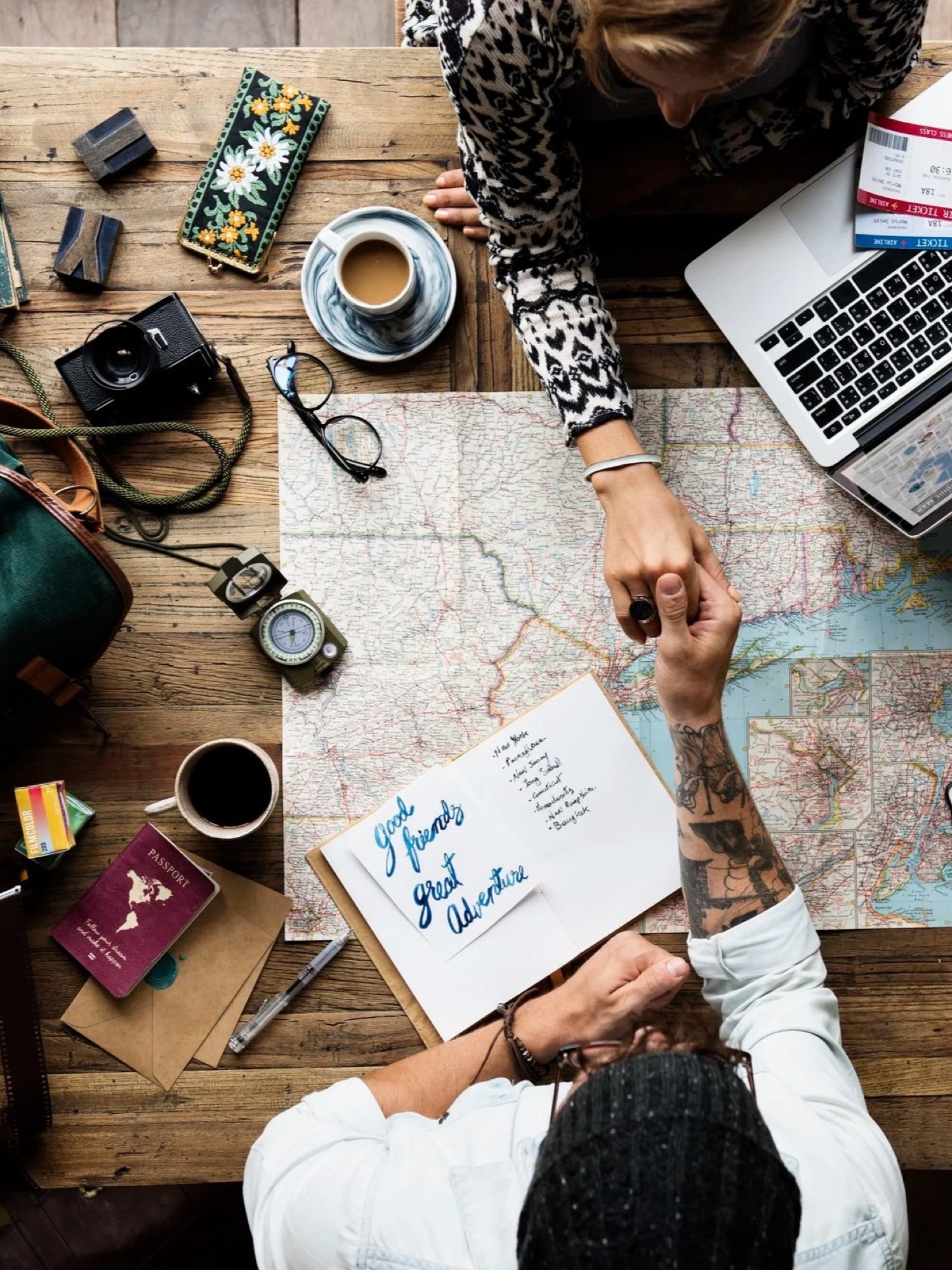 A top-down view of a table covered with maps and travel items, two people holding hands over it.