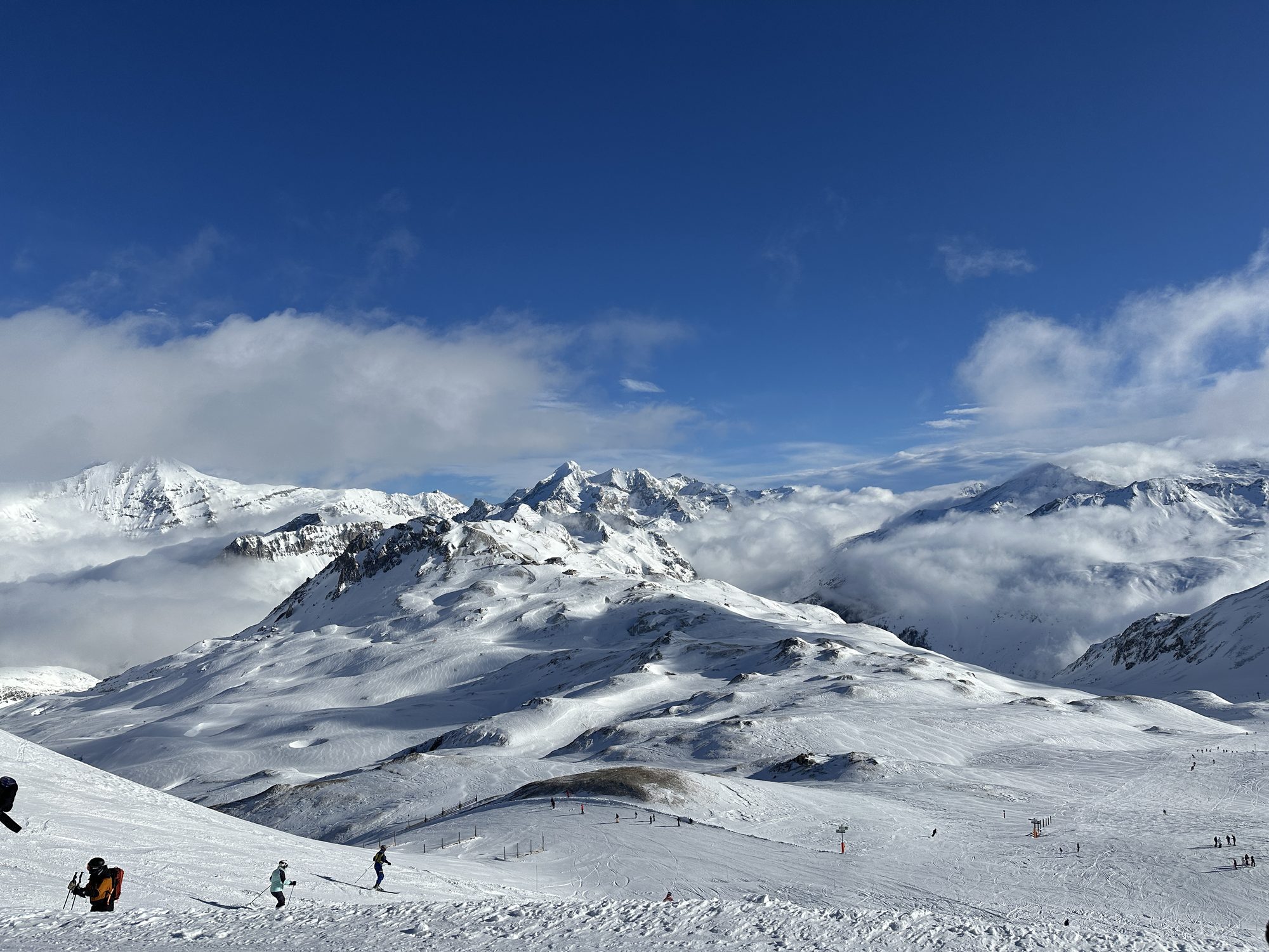 Snow-covered Alpine peaks rising above a sea of cloud, with skiers on a wide groomed piste in the foreground.