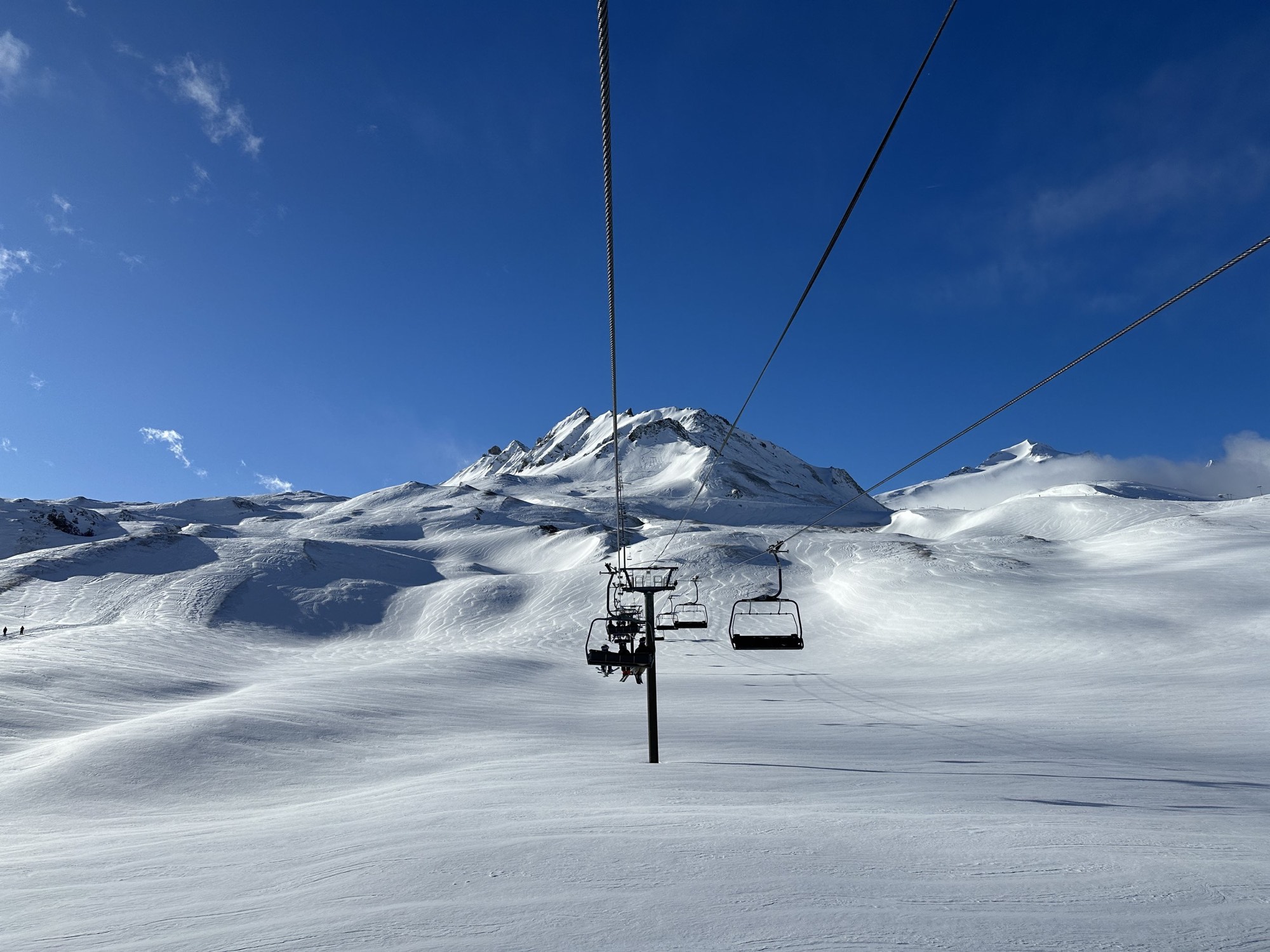 A chairlift climbing above a wide snow-covered alpine bowl under a clear blue sky.