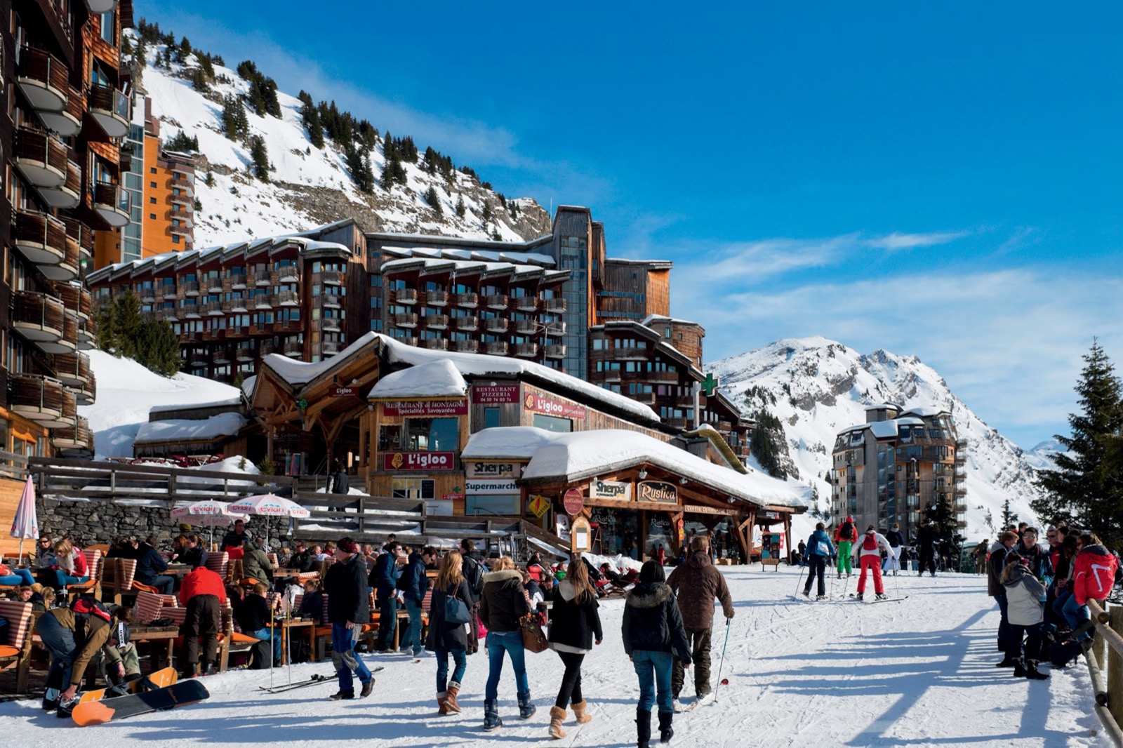 Avoriaz village on a sunny day: skiers and families walking along the snow-covered main street past chalet-style buildings and a busy café terrace.