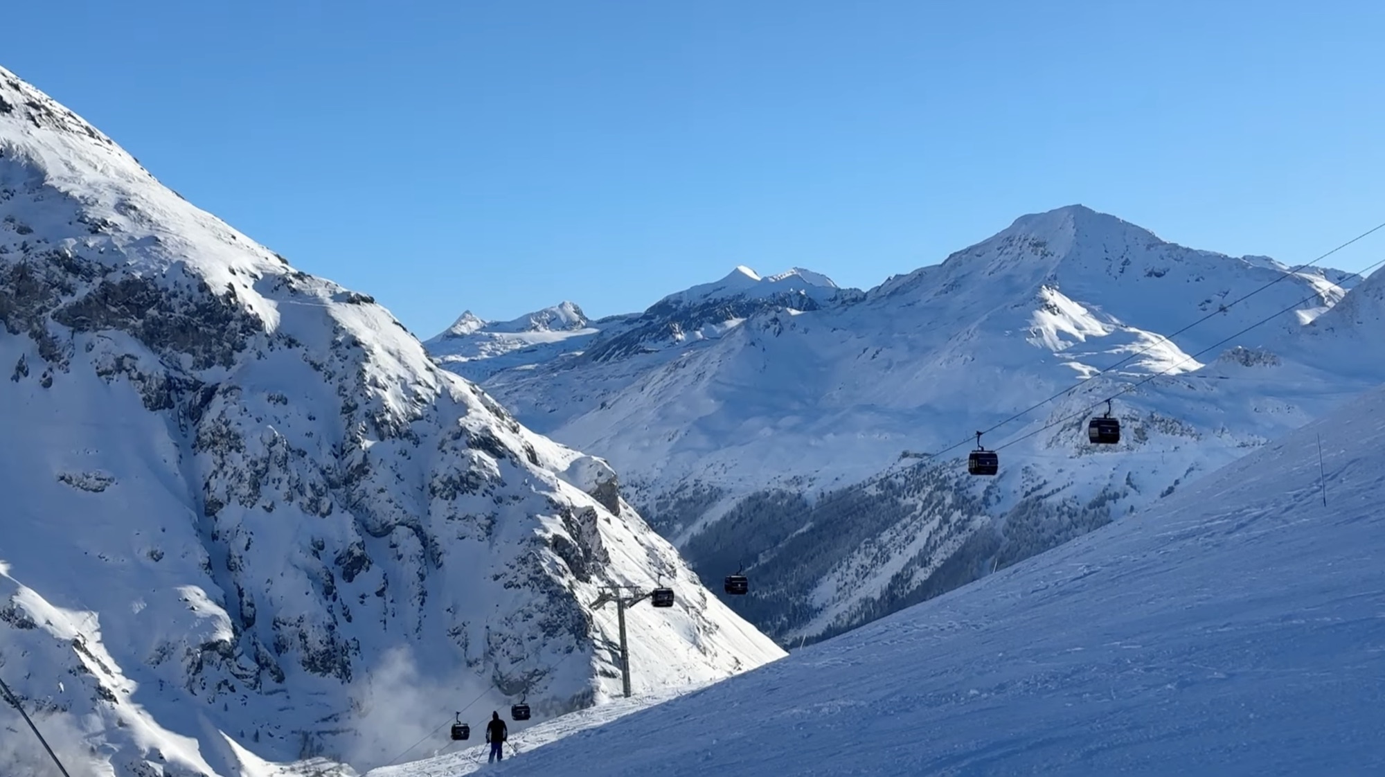 Gondolas running up a snowy valley under a clear blue sky, with a skier on the groomed piste below.